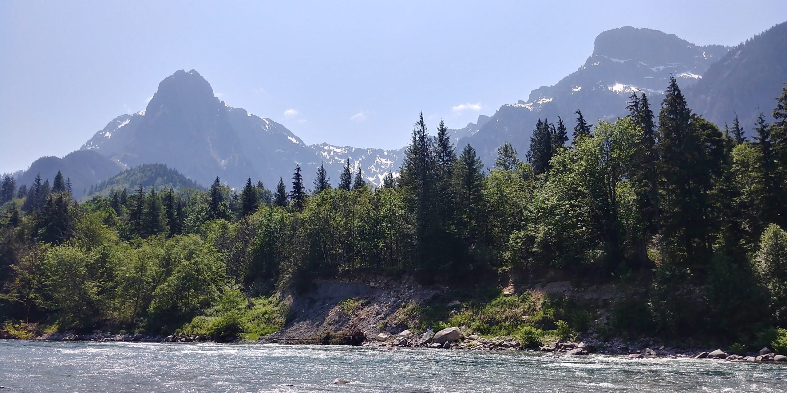 A view of two mountains with some patches of snow and a blue sky. A mix of evergreen and deciduous trees in the foreground.