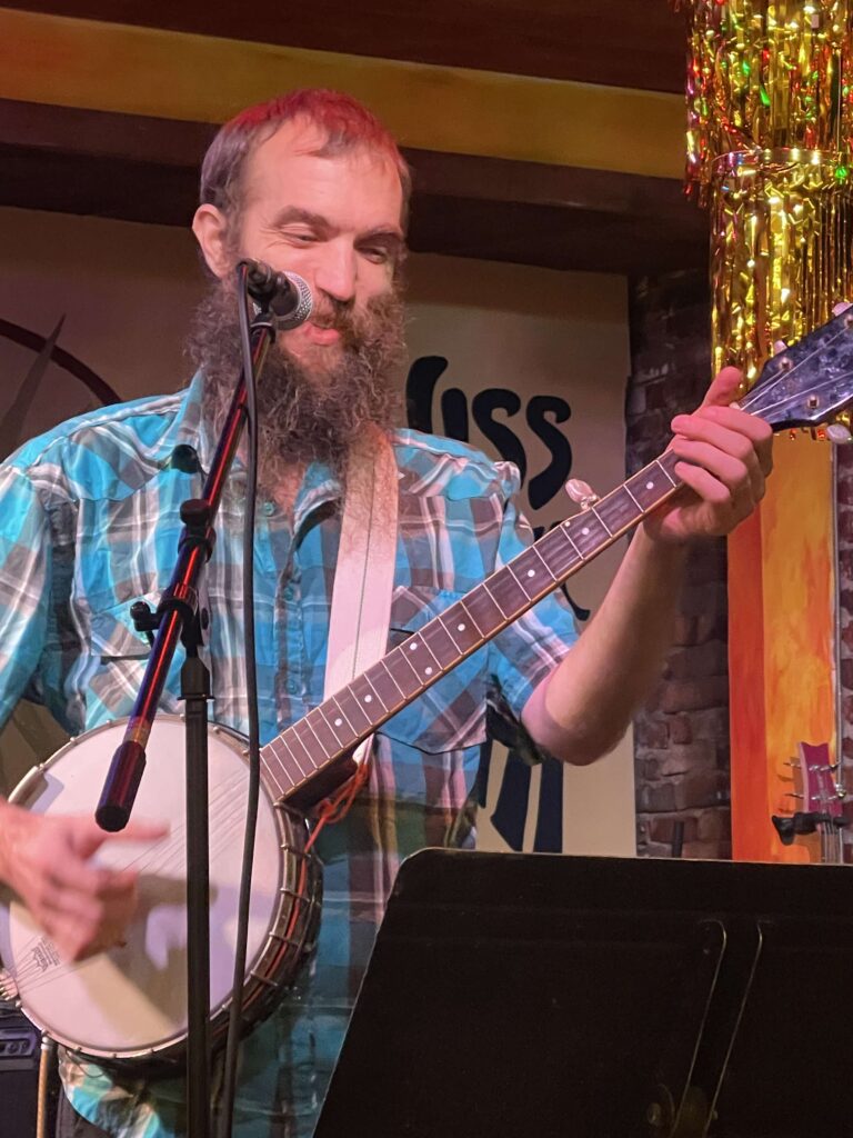 Brian smiling while playing banjo. Brian has short hair and a full beard. He's wearing a blue, gray, and white checked short sleeve shirt.